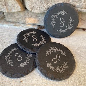 May include: Four round, dark gray slate coasters. Each coaster features a white floral wreath design around a capital letter "S". The coasters are arranged on a light-colored surface, with a stone wall in the background.