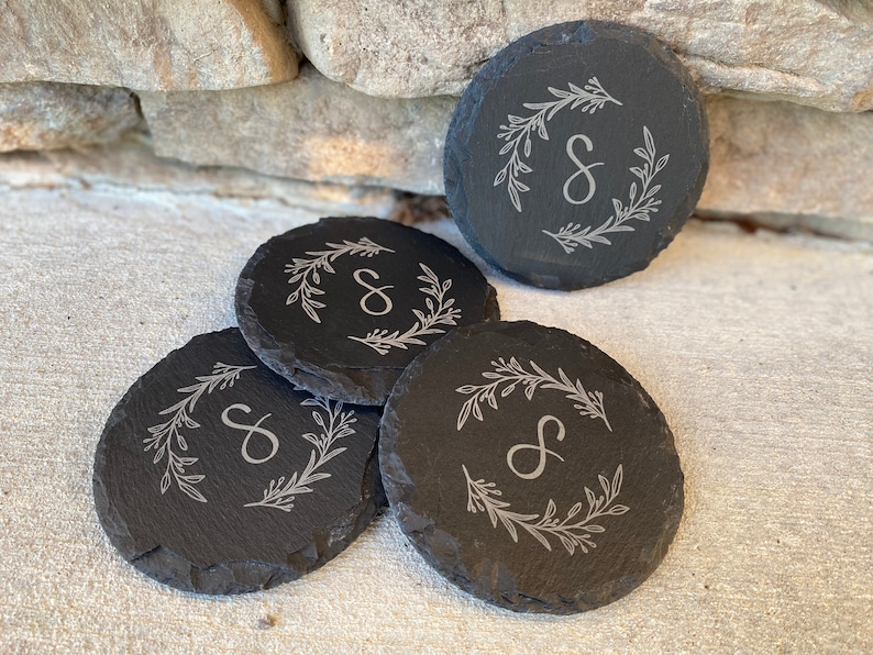May include: Four round, dark gray slate coasters. Each coaster features a white floral wreath design around a capital letter "S". The coasters are arranged on a light-colored surface, with a stone wall in the background.