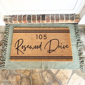 May include: A brown coir doormat with a green border and fringe. The doormat has black text that reads "105 Rosewood Drive" with two black lines above and below the text.