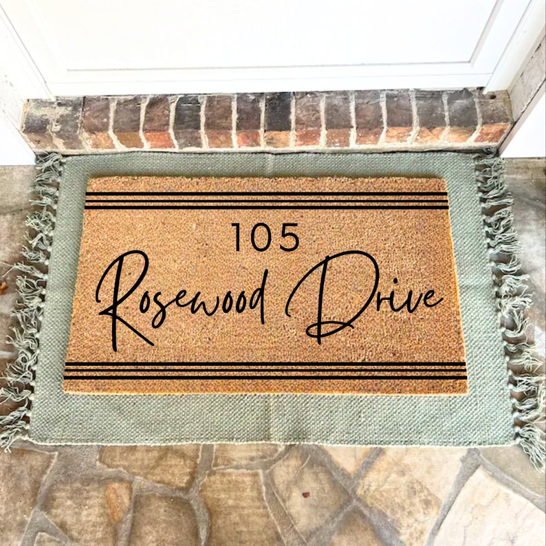 May include: A brown coir doormat with a green border and fringe. The doormat has black text that reads "105 Rosewood Drive" with two black lines above and below the text.