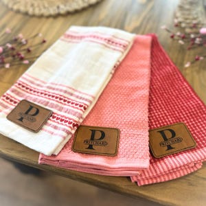May include: Three folded kitchen towels in shades of red and pink, with a white towel featuring red heart and stripe patterns. Each towel has a brown leather patch with the letter "P" and the name "PRITCHARD".