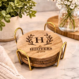May include: A set of four round wooden coasters with a gold-colored metal holder. Each coaster is engraved with the letter "H", laurel branches, and the text "the HOOVERS est. 2026".