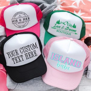 May include: Four trucker hats with white fronts and mesh backs. One is pink with "HIS & HER DECOR" text, another green with "Love & Dreams BOUTIQUE", a black hat with "YOUR CUSTOM TEXT HERE", and a pink hat with "ISLAND babe".