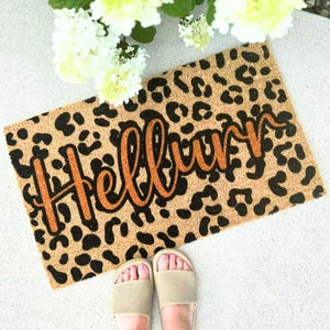 May include: A rectangular doormat with a leopard print background and the word "Hello" in orange script. The mat is made of a natural fiber material and is placed on a light-colored surface. The image also shows a pair of feet wearing beige sandals.