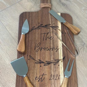 May include: Wooden cheese board with a handle and a set of cheese knives and forks. The board is engraved with "The Browns est. 2026" and a decorative leaf design. The cheese tools have wooden handles and stainless steel blades.