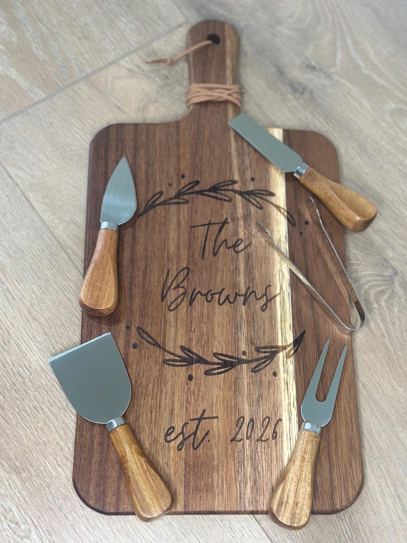 May include: Wooden cheese board with a handle and a set of cheese knives and forks. The board is engraved with "The Browns est. 2026" and a decorative leaf design. The cheese tools have wooden handles and stainless steel blades.