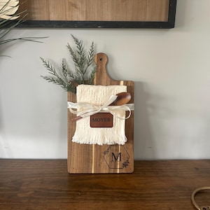 May include: Wooden cutting board with a handle, featuring a cream-colored cloth, wooden spoon, and a leather patch that says "MOYER". The board is decorated with a ribbon and a floral initial "M".