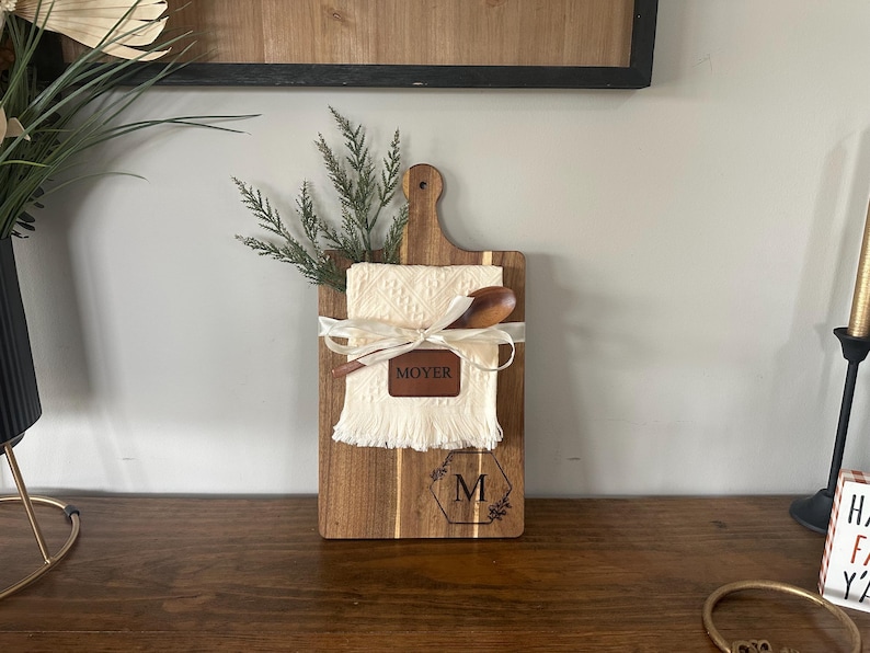 May include: Wooden cutting board with a handle, featuring a cream-colored cloth, wooden spoon, and a leather patch that says "MOYER". The board is decorated with a ribbon and a floral initial "M".