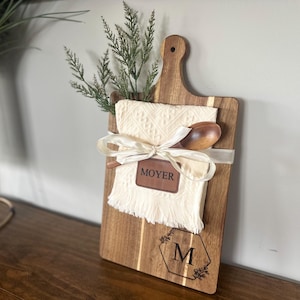 May include: Wooden cutting board with a handle, featuring a cream-colored cloth, wooden spoon, and greenery. A ribbon and a leather tag with the name "MOYER" are tied around the cloth. The board has a decorative "M" and floral design.