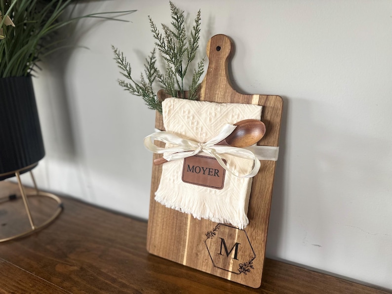 May include: Wooden cutting board with a handle, featuring a cream-colored cloth, wooden spoon, and greenery. A ribbon and a leather tag with the name "MOYER" are tied around the cloth. The board has a decorative "M" and floral design.