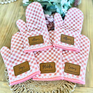 May include: A set of five oven mitts with a pink and white gingham pattern. Each mitt has a brown leatherette patch with a name and "BAKING CO." printed on it. The mitts are trimmed with pink fabric and lace.
