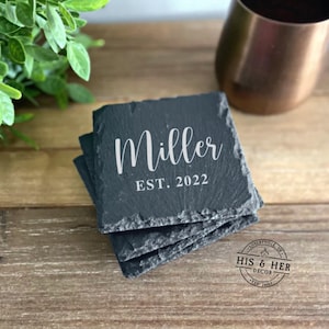 May include: A stack of square, dark gray slate coasters. The coasters are engraved with the name "Miller" in a white script font, with "EST. 2022" below. The coasters are on a wooden surface.