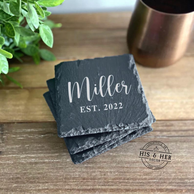 May include: A stack of square, dark gray slate coasters. The coasters are engraved with the name "Miller" in a white script font, with "EST. 2022" below. The coasters are on a wooden surface.