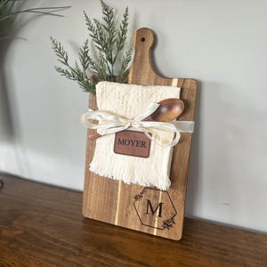 May include: A wooden cutting board with a handle, featuring a cream-colored cloth, wooden spoon, and greenery. The board is adorned with a leather tag that reads "MOYER" and a monogram with the letter "M".