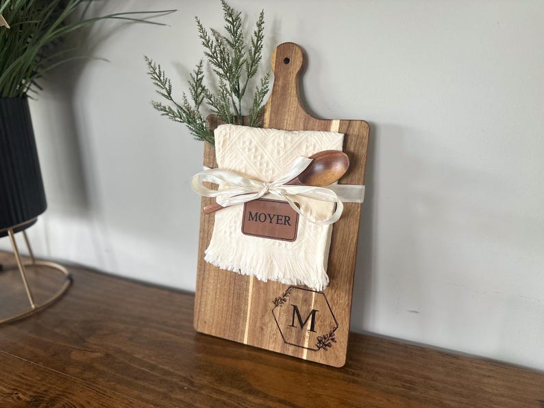 May include: A wooden cutting board with a handle, featuring a cream-colored cloth, wooden spoon, and greenery. The board is adorned with a leather tag that reads "MOYER" and a monogram with the letter "M".