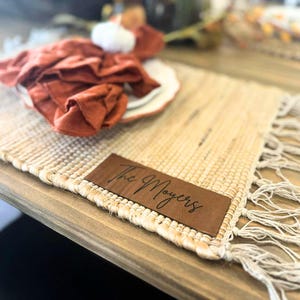 May include: A woven placemat with a fringed edge and a brown leather patch that reads "The Moyers" in script. A rust-colored napkin and a white plate with an orange rim are on top of the placemat. The placemat is on a wooden table.