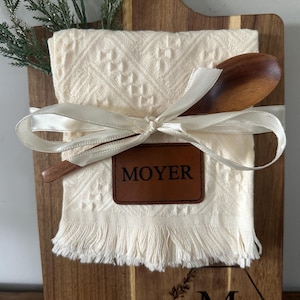 May include: A wooden cutting board with a cream-colored kitchen towel, wooden spoon, and a leather tag that reads "MOYER". The towel is tied with a white ribbon and has a fringed edge. A sprig of greenery is on the left.