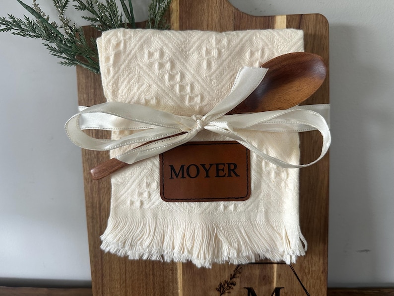 May include: A wooden cutting board with a cream-colored kitchen towel, wooden spoon, and a leather tag that reads "MOYER". The towel is tied with a white ribbon and has a fringed edge. A sprig of greenery is on the left.