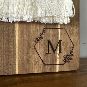 May include: Wooden cutting board with a decorative hexagon design and the letter "M" engraved in the center. The hexagon is framed by floral elements. The top of the board has a white, fringed textile.