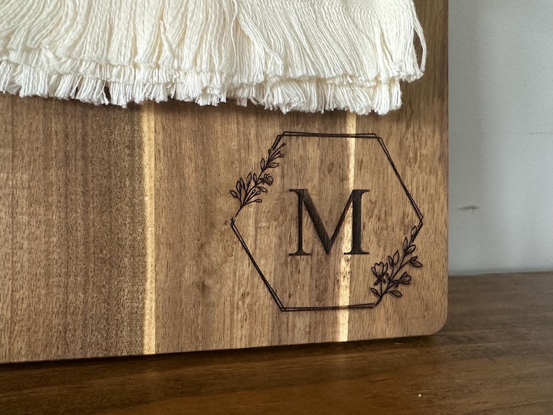May include: Wooden cutting board with a decorative hexagon design and the letter "M" engraved in the center. The hexagon is framed by floral elements. The top of the board has a white, fringed textile.