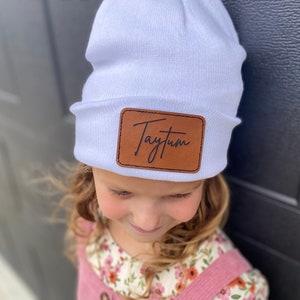 May include: White beanie with a brown leather patch that says "Taytum" in cursive.