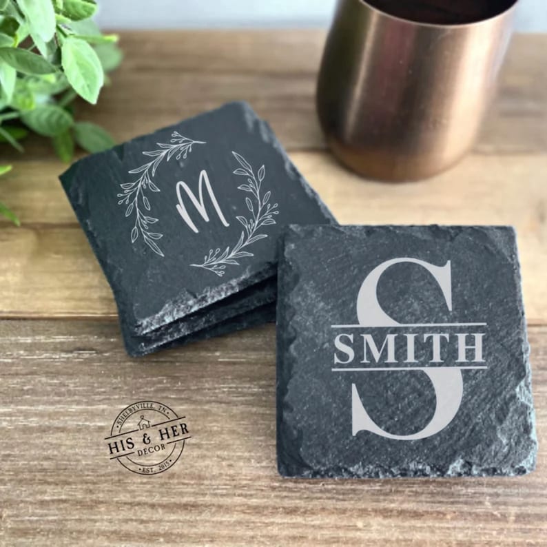 May include: Set of slate coasters with personalized designs. One coaster features the letter "M" inside a floral wreath, while another displays "SMITH" with a large "S". The coasters are dark gray and sit on a wooden surface.