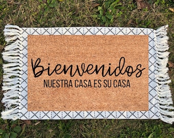 May include: A brown coir doormat with white fringe and a black and white diamond pattern border. The text "Bienvenidos" and "Nuestra casa es su casa" is printed in black on the doormat.