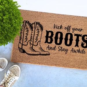 May include: A rectangular doormat with a brown surface features a black illustration of two cowboy boots. The text "Kick off your BOOTS And Stay Awhile" is printed in black, inviting guests to relax.