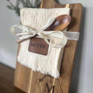 May include: A wooden serving board with a handle, a cream-colored cloth, and a wooden spoon tied with a white ribbon. The board has a leather patch that reads "MOYER" and a monogram "M" with floral accents.