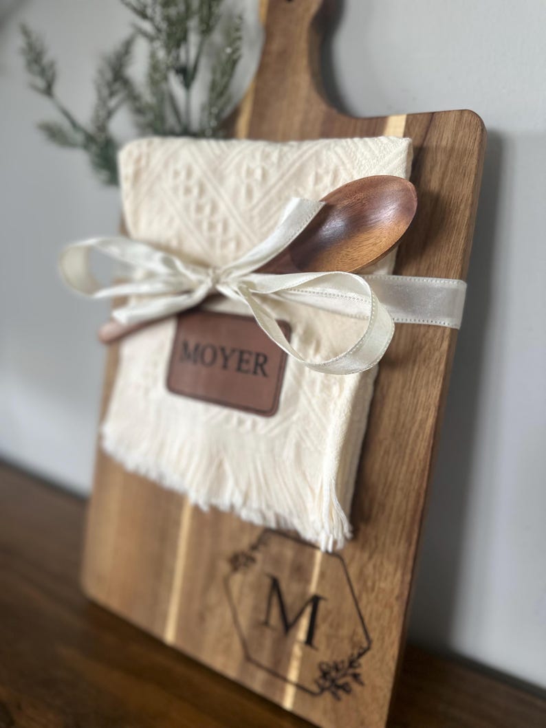 May include: A wooden serving board with a handle, a cream-colored cloth, and a wooden spoon tied with a white ribbon. The board has a leather patch that reads "MOYER" and a monogram "M" with floral accents.