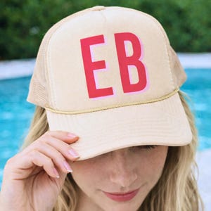 May include: Tan trucker hat with a mesh back and a curved brim. The hat features the letters "EB" in a bold, pink font on the front panel. A gold-colored rope detail accents the brim.