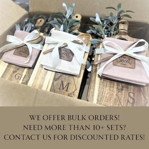 May include: Wooden cutting boards with engraved initials and decorative floral designs are paired with pink waffle weave towels, wooden spoons, and tied with white ribbons. The words "Sweet Home" are on a leather patch. The items are in a cardboard box.
