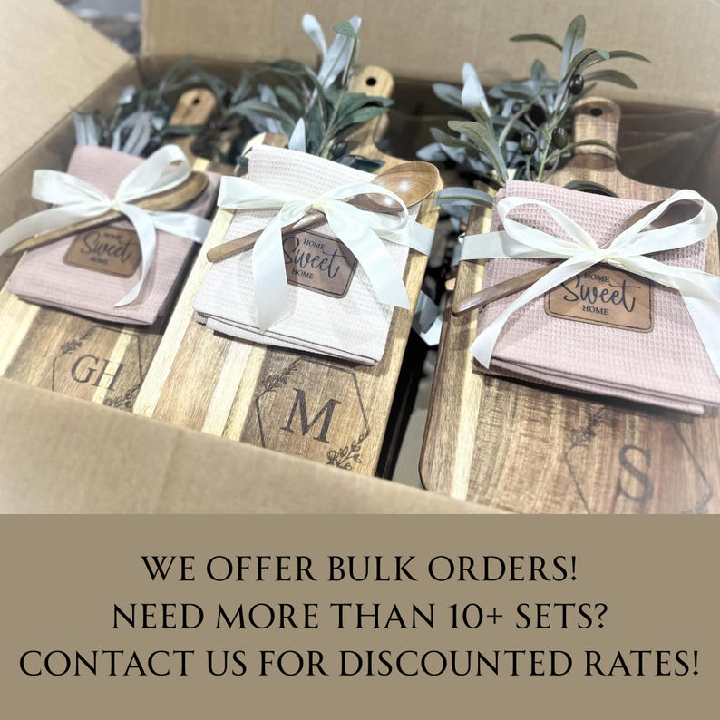 May include: Wooden cutting boards with engraved initials and decorative floral designs are paired with pink waffle weave towels, wooden spoons, and tied with white ribbons. The words "Sweet Home" are on a leather patch. The items are in a cardboard box.