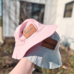 May include: Two bucket hats, one pink and one light blue, with brown leather patches. The pink hat has the name "Tayluhn" on the patch and the blue hat has the name "LINCOLN" on the patch.