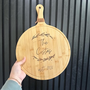 May include: Round wooden cutting board with a handle, featuring the engraved text "The Costas" and "est. 2026" surrounded by leaf designs. The board is light brown and has a natural wood grain.