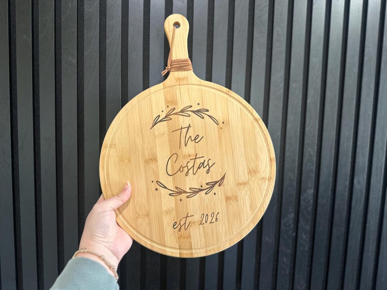 May include: Round wooden cutting board with a handle, featuring the engraved text "The Costas" and "est. 2026" surrounded by leaf designs. The board is light brown and has a natural wood grain.