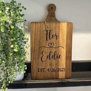 May include: A wooden cutting board with a handle and a decorative string. The board is engraved with the names "Flor" and "Eddie", a heart symbol, and the date "EST. 4/26/2025". The board is displayed on a black shelf.