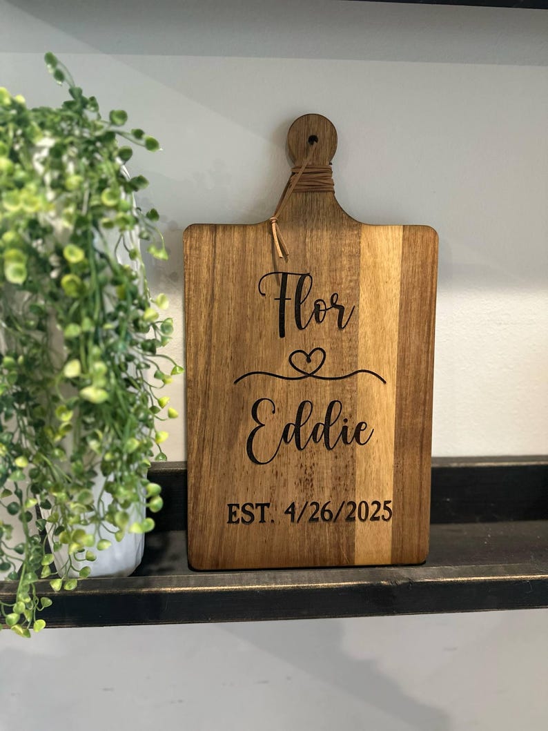 May include: A wooden cutting board with a handle and a decorative string. The board is engraved with the names "Flor" and "Eddie", a heart symbol, and the date "EST. 4/26/2025". The board is displayed on a black shelf.