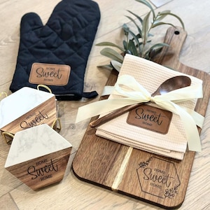 May include: A kitchen-themed gift set featuring a black oven mitt, marble and wood coasters, a wooden cutting board, a beige waffle-weave towel, and a wooden spoon. Each item has a brown leatherette tag with the words "Home Sweet Home".