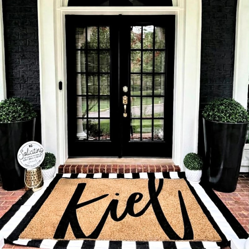 Puede incluir: Una puerta principal negra con puertas dobles y molduras blancas. Una alfombra de bienvenida beige con la palabra "Kiel" en escritura negra, enmarcada por rayas blancas y negras. Las jardineras negras con vegetaci&oacute;n flanquean la puerta, con un letrero de "No Soliciting".