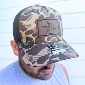 May include: Camouflage pattern Richardson 112 trucker hat with a brown leather patch. The patch features a fist bump design with the names "Walker", "Eleanor", "Daddy", and "Maverick" and the text "Since 2019". Adjustable fit.