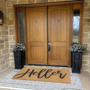 Puede incluir: Una entrada acogedora con puertas dobles de madera, jardineras negras con vegetaci&oacute;n a ambos lados. Una alfombra con la palabra "Hello" en escritura se encuentra sobre una alfombra a rayas blancas y negras. La puerta es de madera.