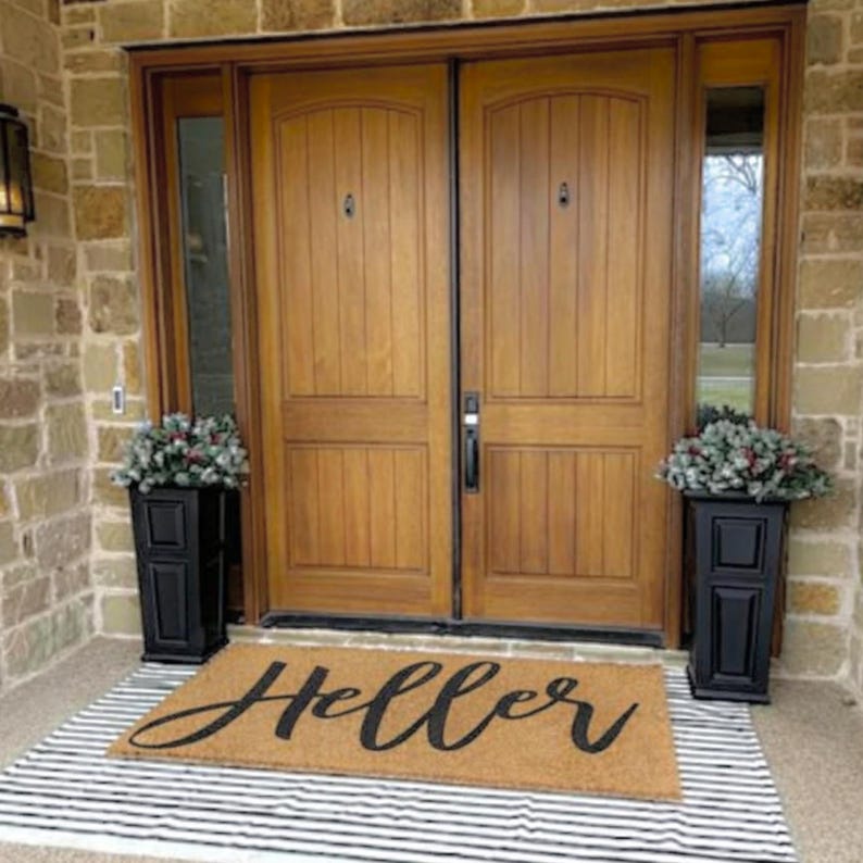 Puede incluir: Una entrada acogedora con puertas dobles de madera, jardineras negras con vegetaci&oacute;n a ambos lados. Una alfombra con la palabra "Hello" en escritura se encuentra sobre una alfombra a rayas blancas y negras. La puerta es de madera.
