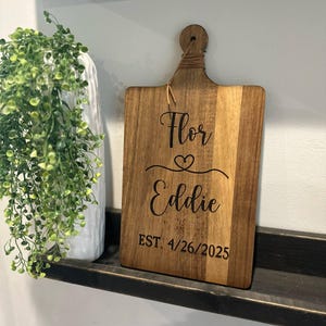 May include: Wooden cutting board with a handle, featuring the names "Flor" and "Eddie" with a heart symbol, and the date "EST. 4/26/2025" engraved in black. The board is made of light and dark brown wood.
