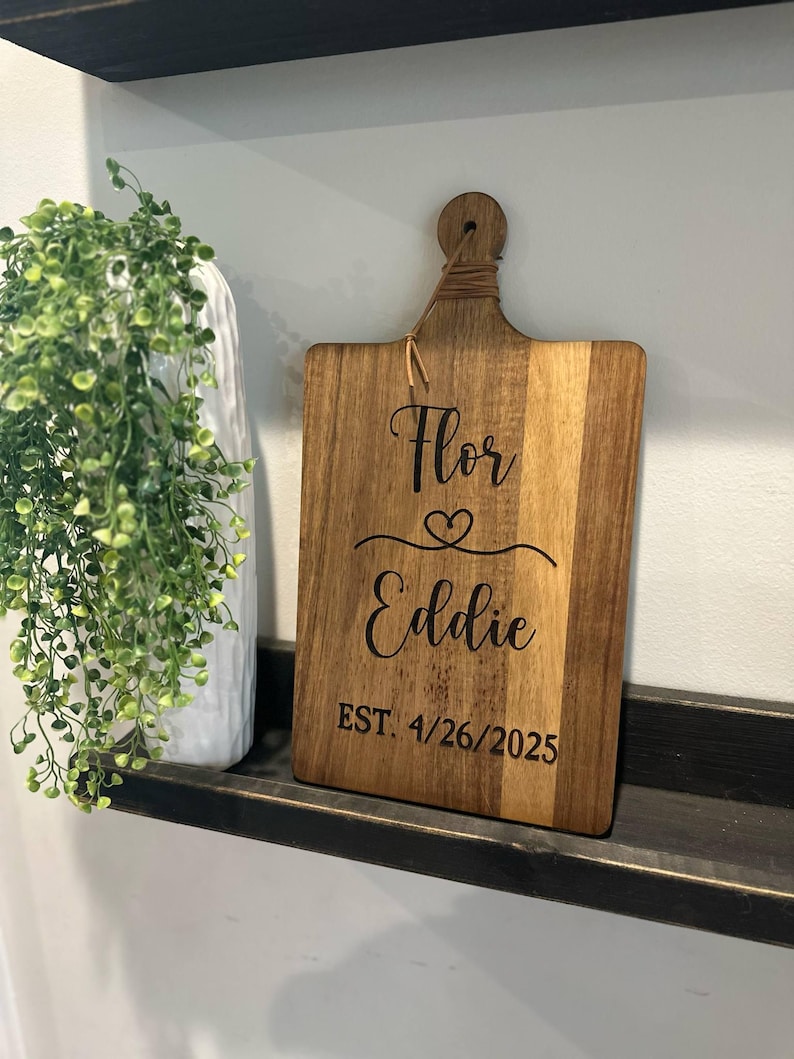 May include: Wooden cutting board with a handle, featuring the names "Flor" and "Eddie" with a heart symbol, and the date "EST. 4/26/2025" engraved in black. The board is made of light and dark brown wood.