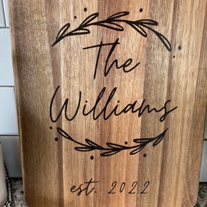 May include: A rectangular wooden cutting board with a light brown finish. The board features the text "The Williams" in a cursive font, encircled by a decorative wreath. Below the name is "est. 2022".
