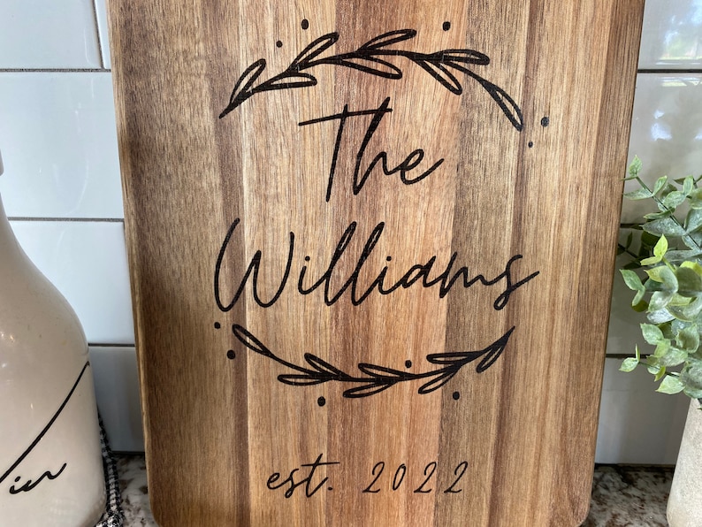 May include: A rectangular wooden cutting board with a light brown finish. The board features the text "The Williams" in a cursive font, encircled by a decorative wreath. Below the name is "est. 2022".