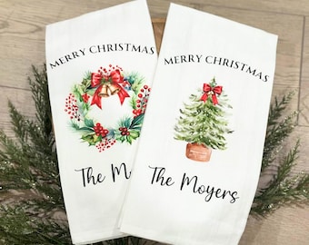 May include: Two white Christmas-themed kitchen towels. One features a wreath with a red bow and bells, the other a Christmas tree with a red bow. Both towels have the text "MERRY CHRISTMAS" and a family name.