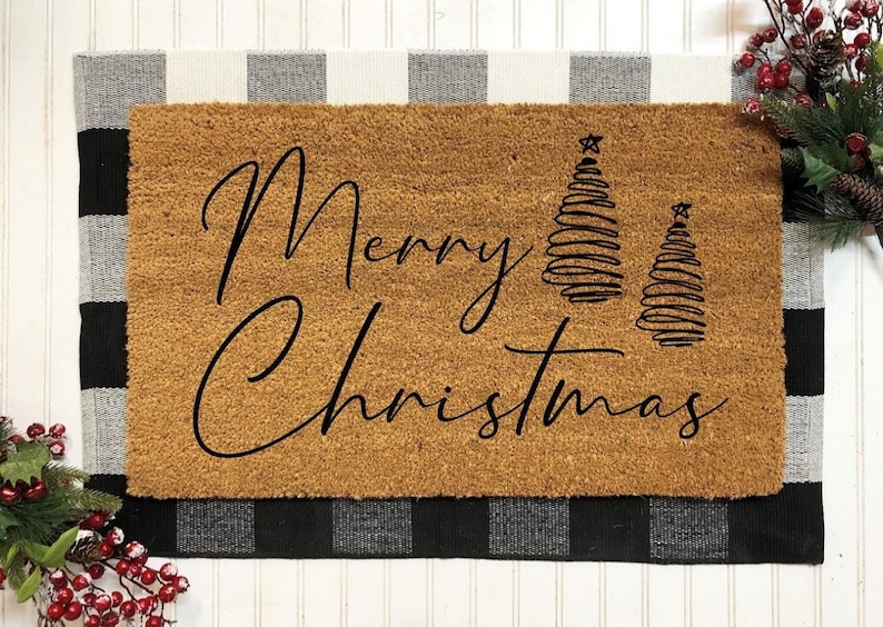 May include: A brown coir doormat with the text "Merry Christmas" in black cursive script. Two stylized Christmas trees are to the right of the text.