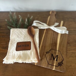 May include: A wooden cutting board with a handle, a white ribbon, and a monogrammed design. A white fringed cloth with a leather patch that says "MOYER" and a wooden spoon are next to the board. A sprig of greenery is also present.